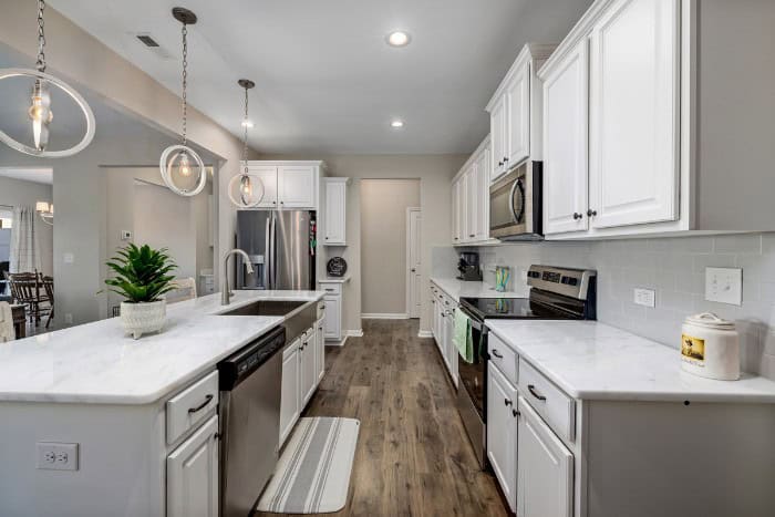 Modern kitchen with white cabinets, stainless steel appliances, marble countertops, and pendant lights above a central island. Wooden flooring and light gray walls complete the space.