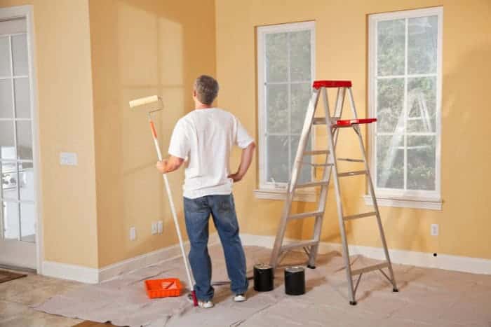 A person stands in a room with light yellow walls, holding a paint roller. A step ladder, paint cans, and painting supplies are visible. The floor is covered with a drop cloth.