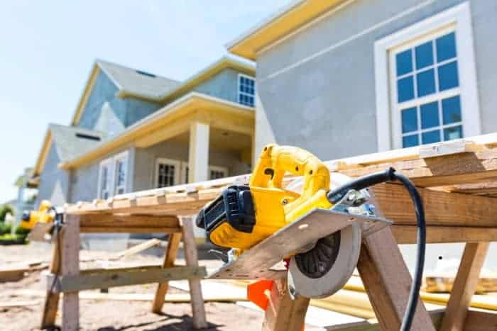 Yellow power saw rests on a makeshift wooden workbench outside a partially constructed house with gray exterior walls and white-framed windows.