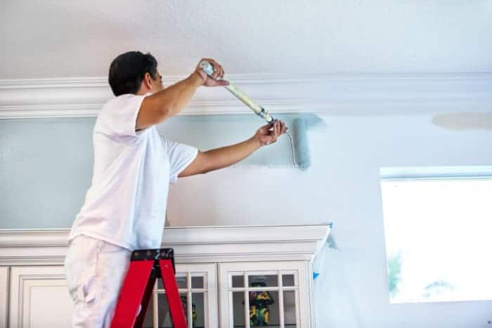 A man standing on a red ladder paints the upper section of a light blue wall near the ceiling using a paint roller.